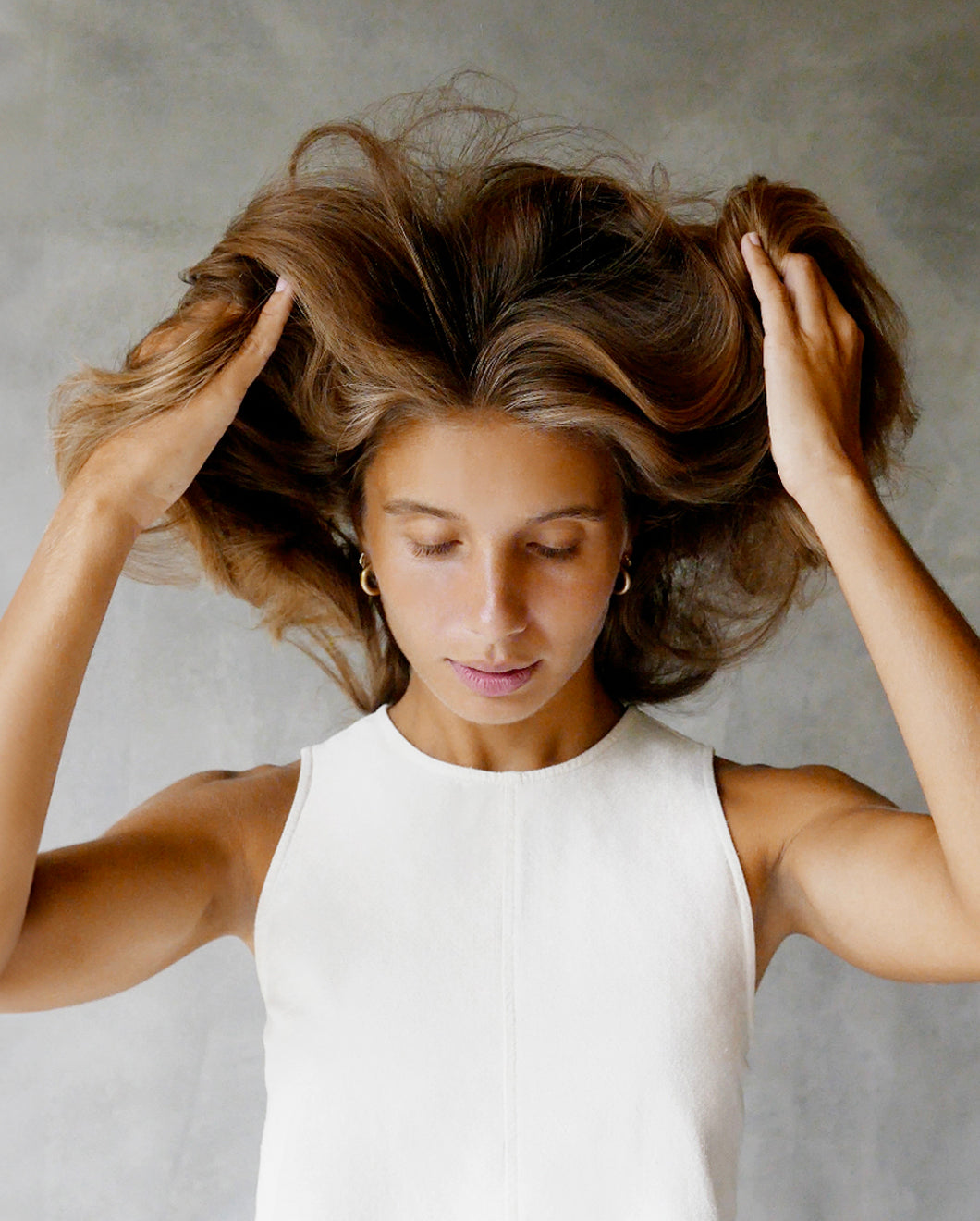 Young woman in a white sleeveless top lifting her voluminous hair with both hands, eyes closed, against a soft gray background.