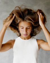 Load image into Gallery viewer, Young woman in a white sleeveless top lifting her voluminous hair with both hands, eyes closed, against a soft gray background.