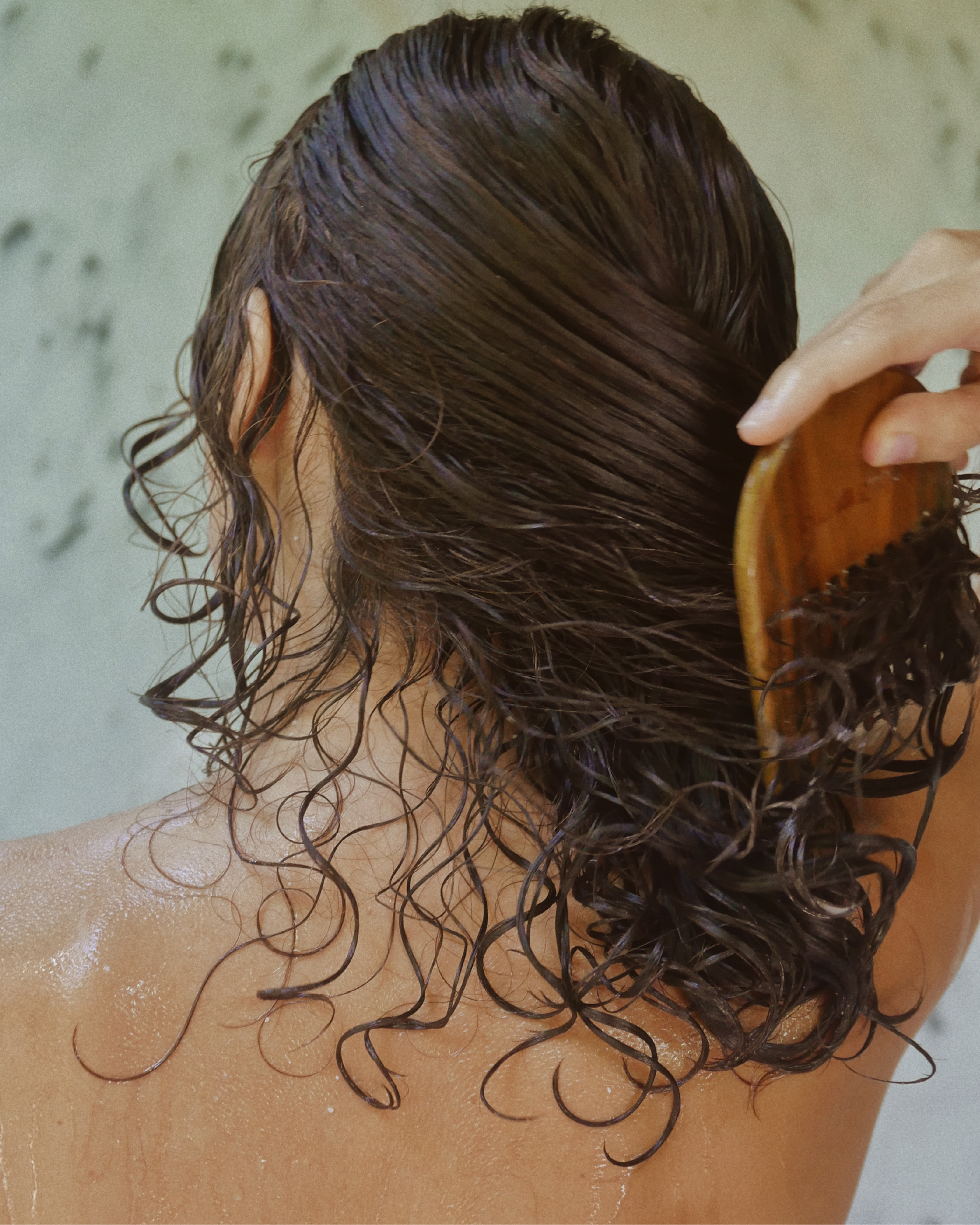 Back view of a person combing wet, wavy hair with a wooden hair brush in a shower setting.