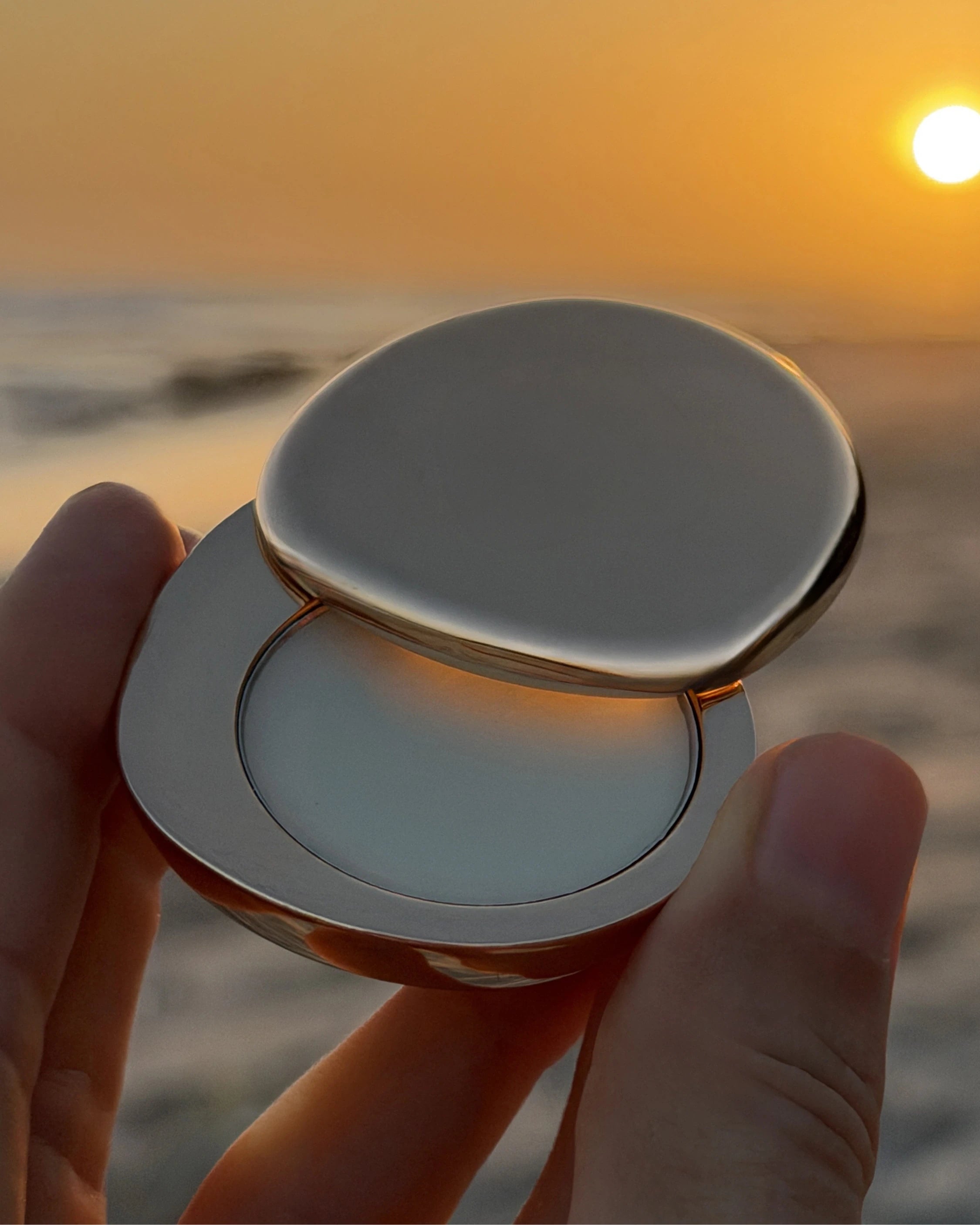 Close-up of Blu & Green's Juniper Dawn Solid Scent, case slightly open, being held in front of a colorful sunrise.