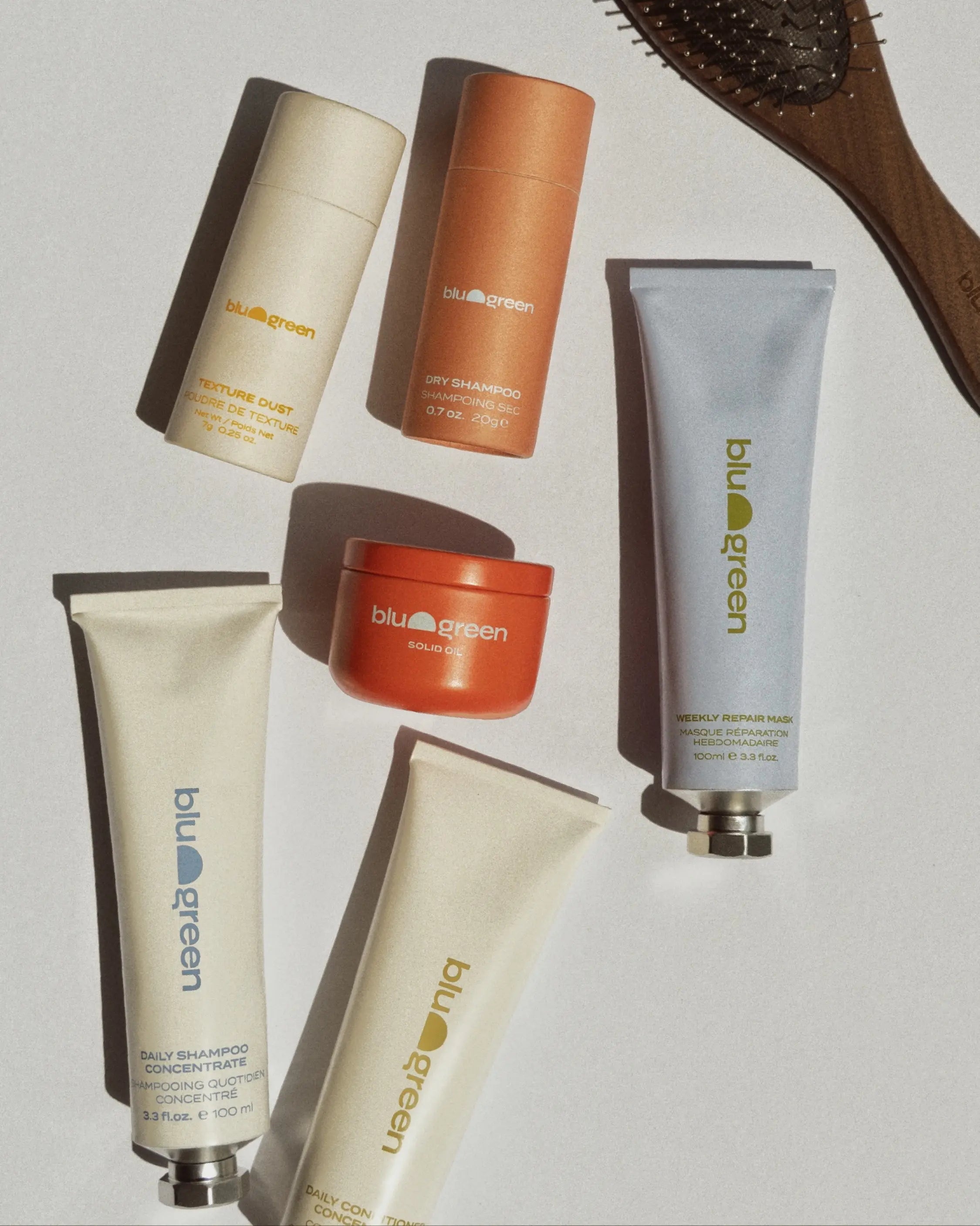 Flat lay of Blu & Green hair care products, including Solid Oil, Texture Dust, Dry Shampoo, Walnut Detangling Brush, Weekly Repair Mask, and Daily Shampoo & Conditioner Concentrates.