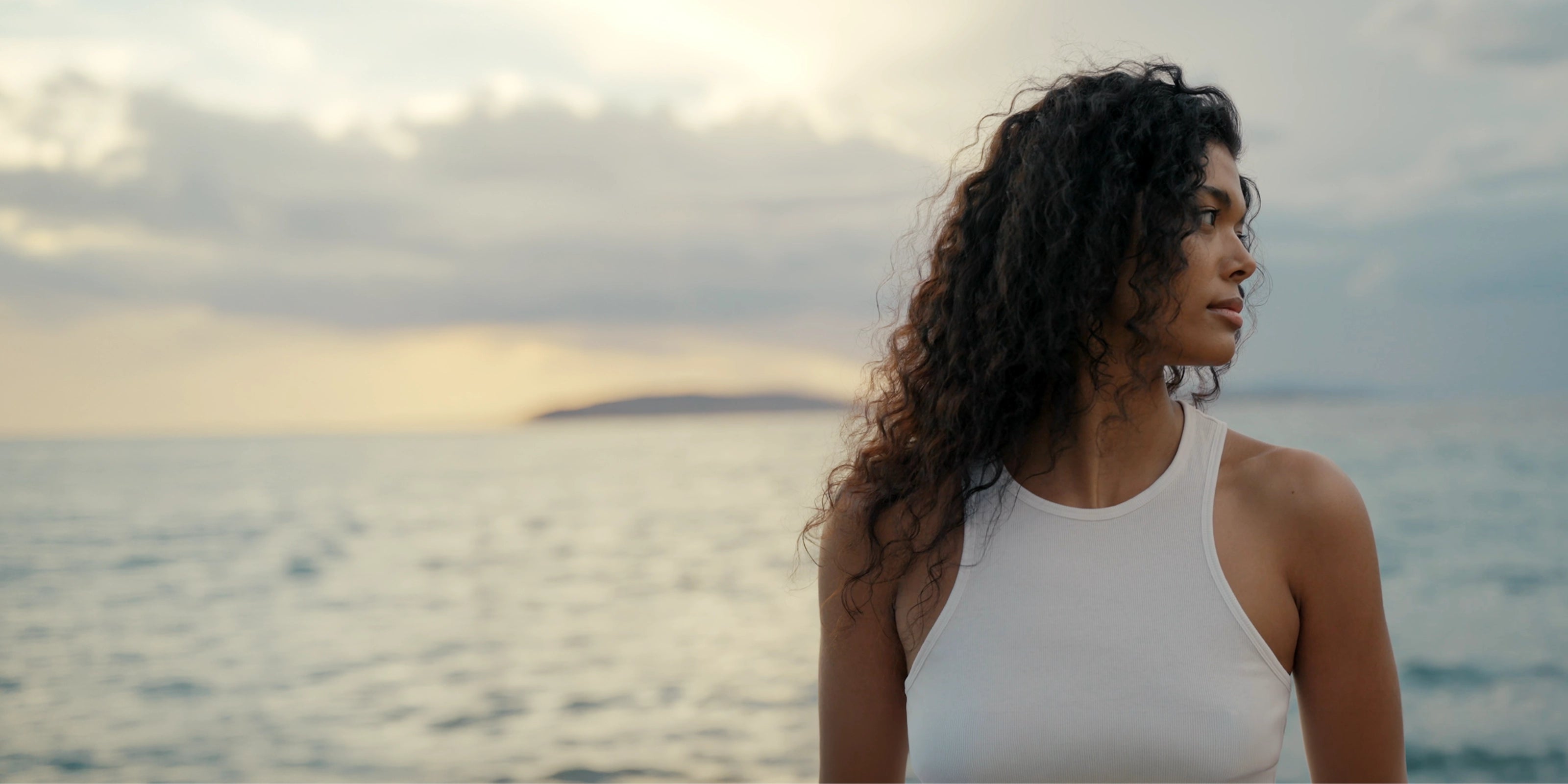 Woman in a white tank top standing by the ocean with a cloudy sky.