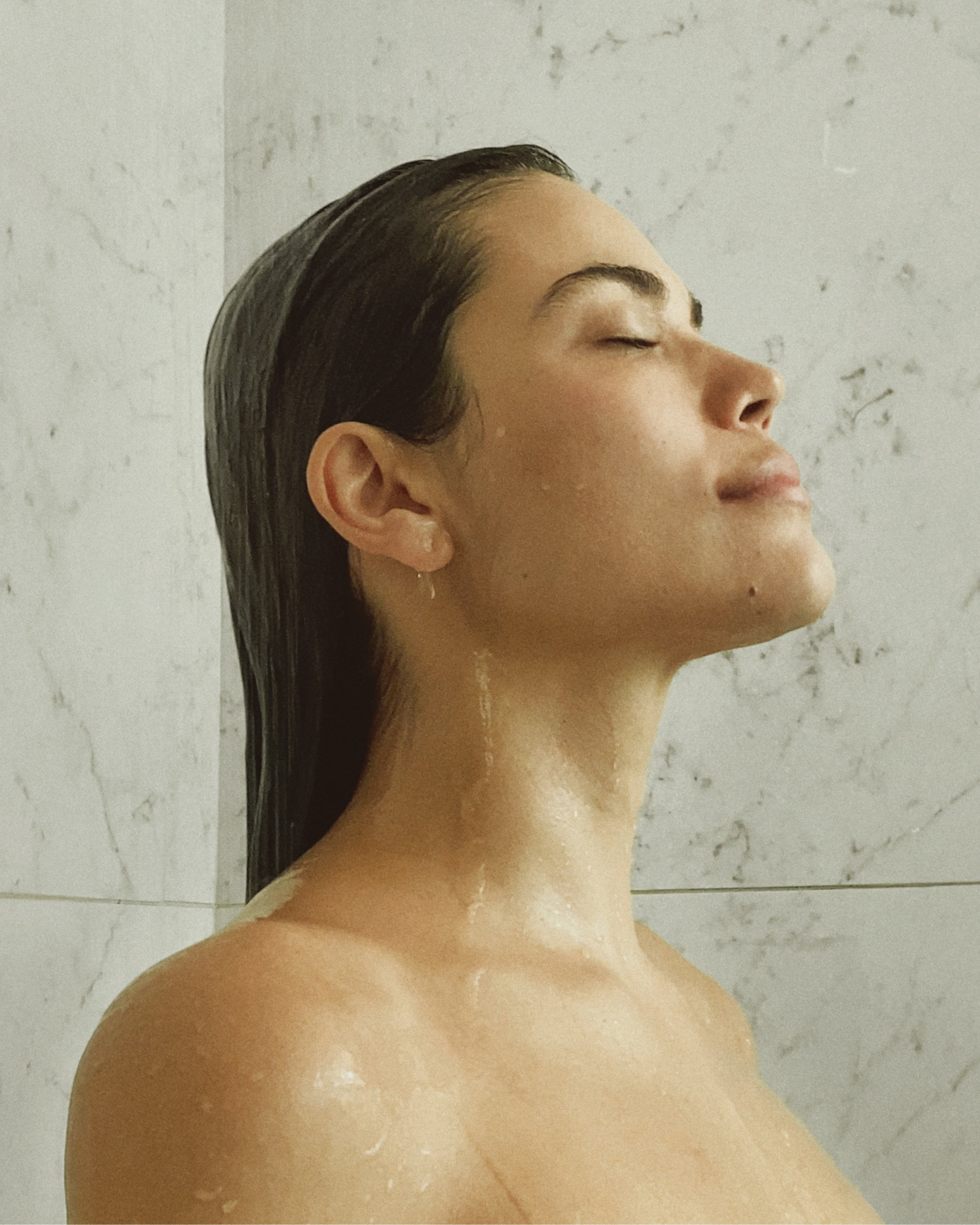 A woman standing under running water with slicked-back hair in a marble shower.