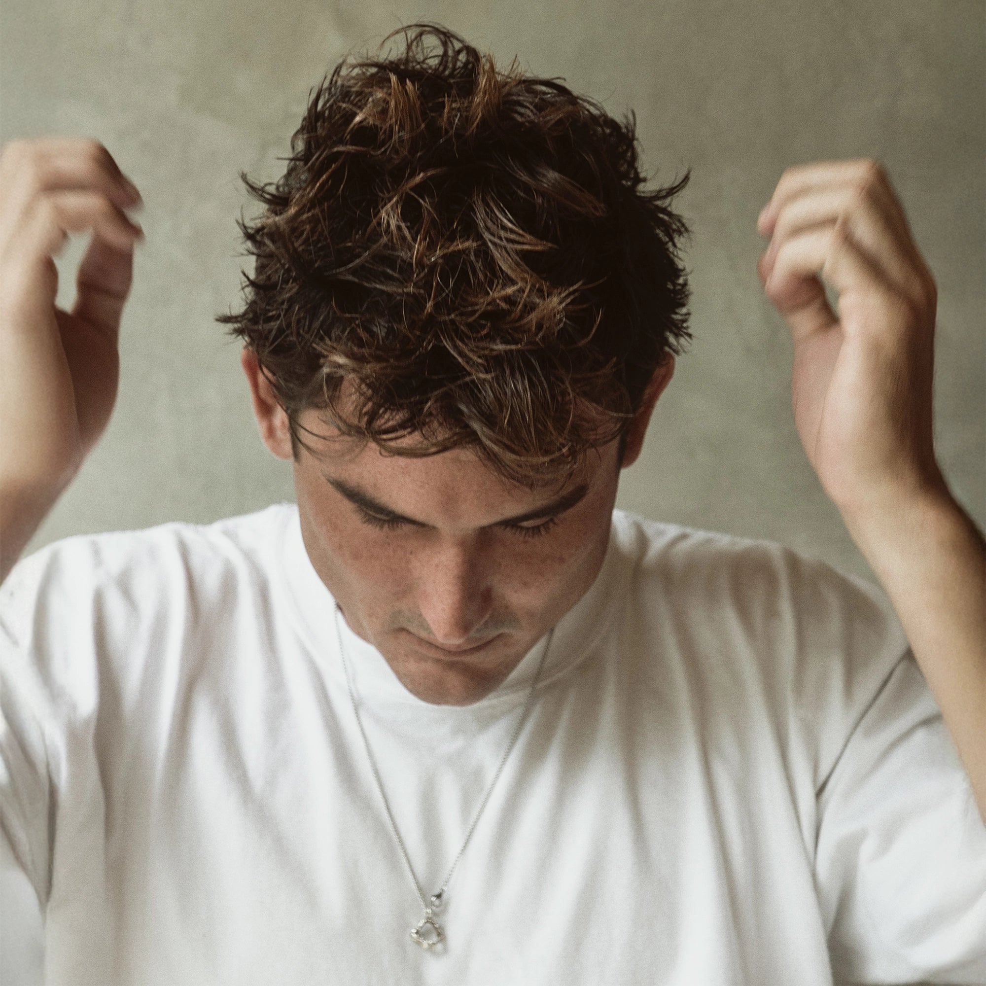 Portrait of a man with tousled short hair looking downward, hands raised near his head.