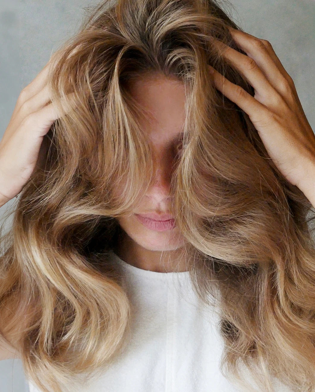 Close-up of a woman running her hands through her voluminous, wavy hair, head tilted down, wearing a white sleeveless top against a neutral grey background.