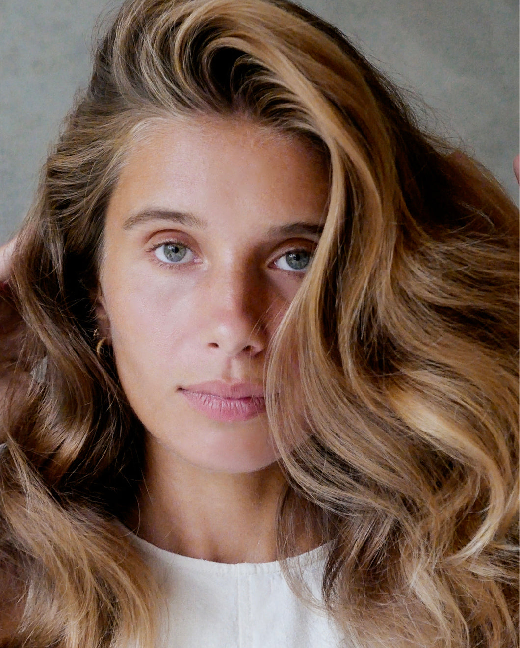 Close-up of a woman with soft, voluminous waves, looking directly at the camera against a neutral grey background.