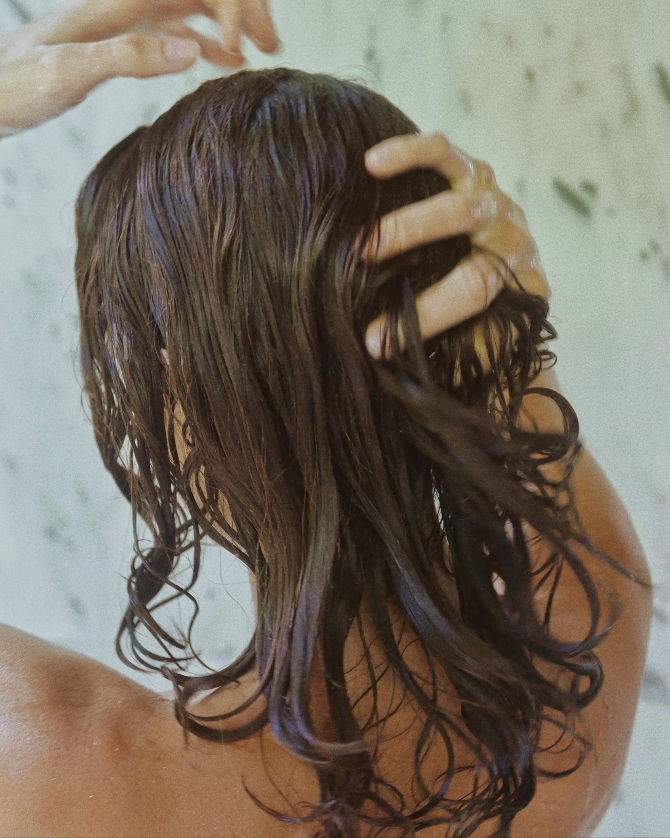 Back view of a person running their hands through wet hair in a marble shower.