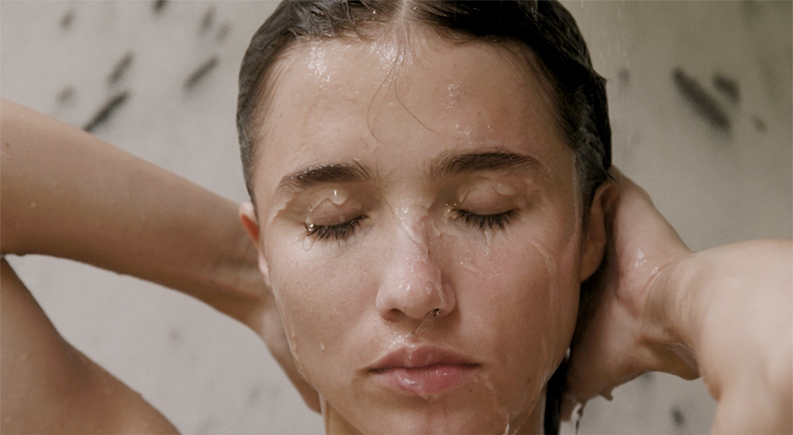 Woman in the shower with wet hair and water droplets on her face against a neutral background