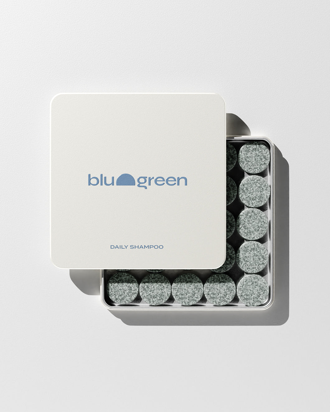 blu and green daily shampoo 50 muted green colored tablets in a flat square tin with blue logo. lid slightly off, studio, white ground, and strong light.