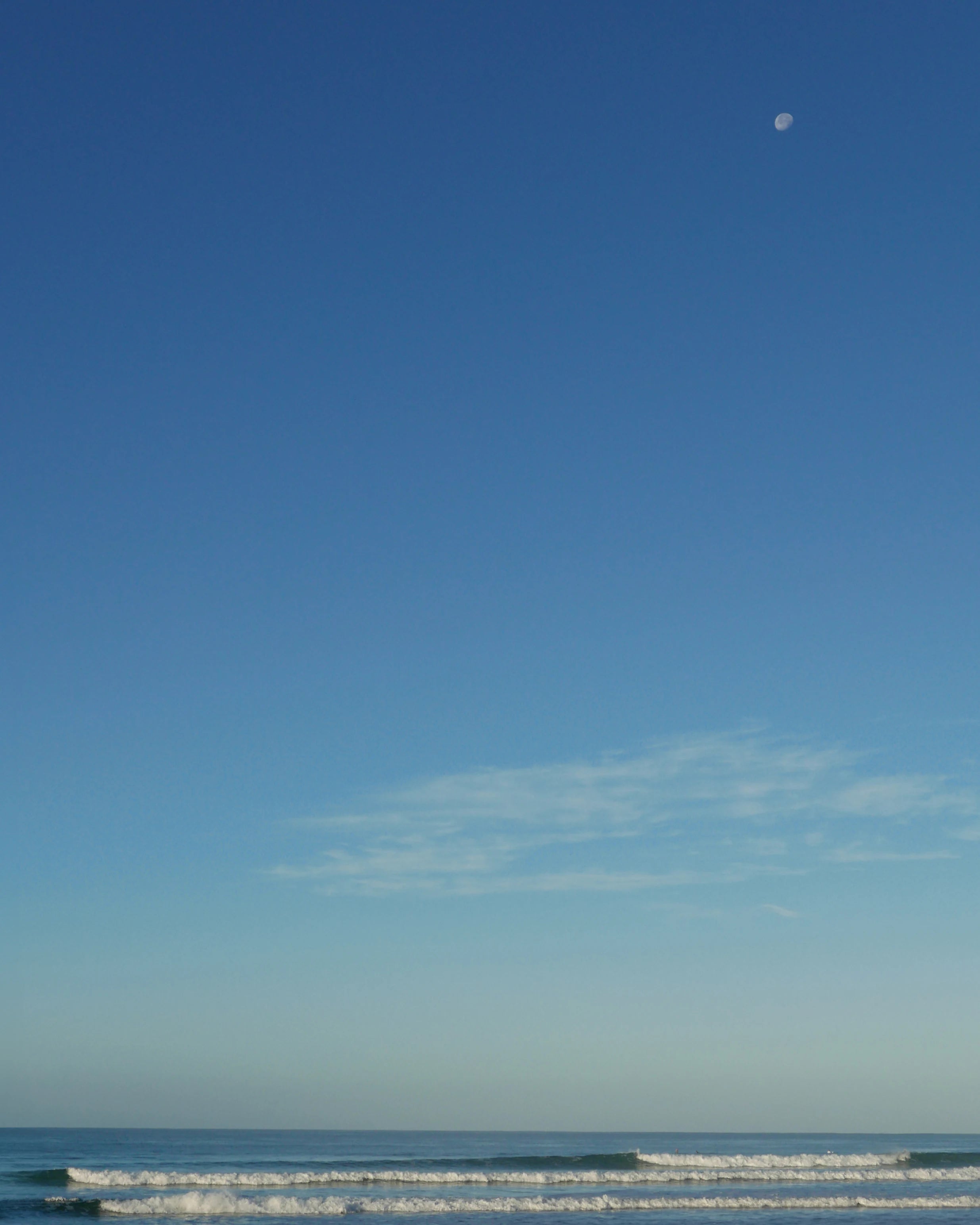 Soft ocean waves with a bright blue sky, subtle clouds, and the moon showing subtly