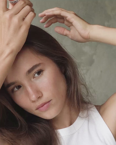 Young woman with brunette hair tapping Blu & Green aerosol-free Dry Shampoo powder on her roots, then working the product through with her fingers.